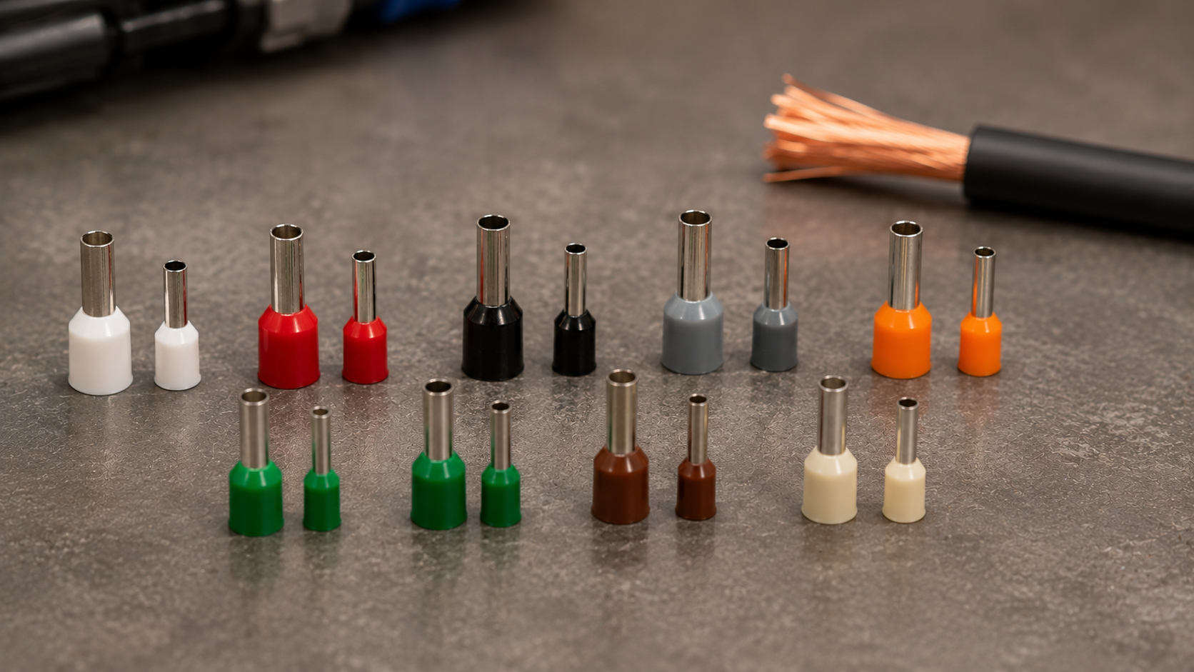 Colour-coded insulated bootlace ferrules in multiple sizes arranged on a workshop bench beside stripped copper wire